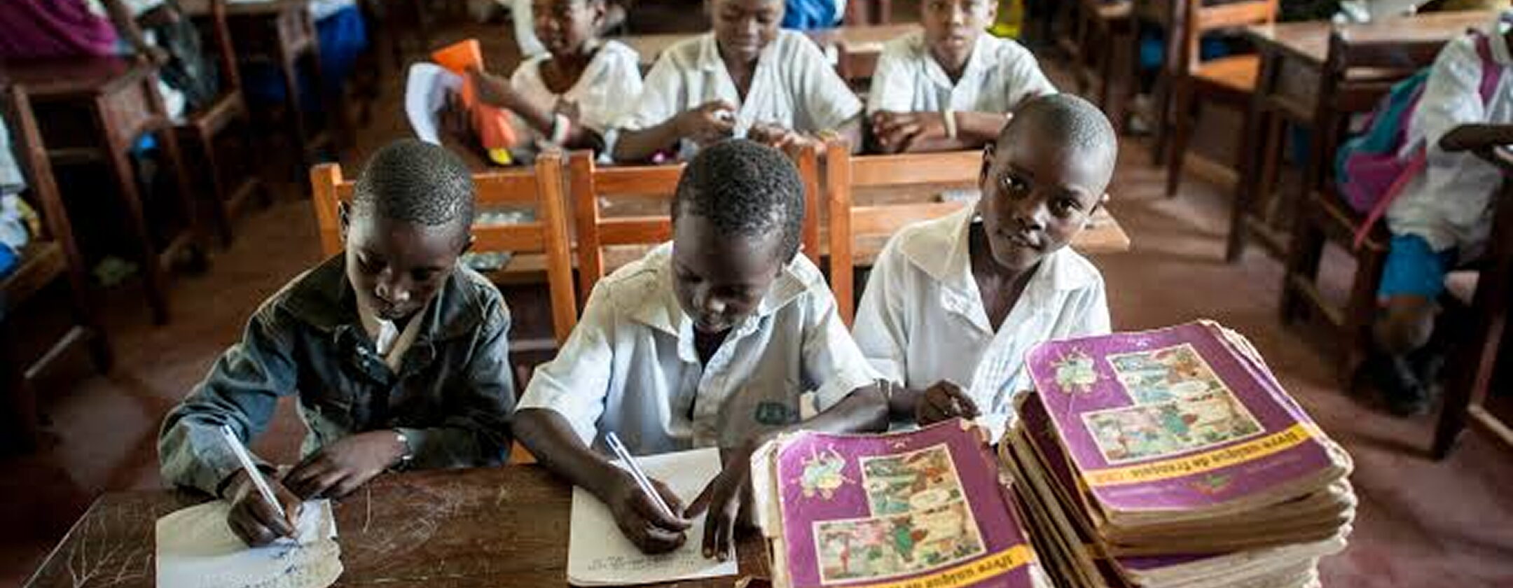 Pupils writing in exercise books at wooden desks in an FGC Kano classroom